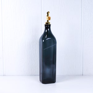 May include: A dark grey glass bottle with a gold metal pump top. The bottle is sitting on a white surface.