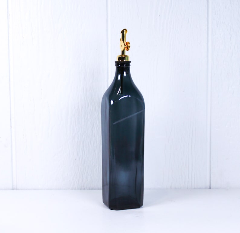 May include: A dark grey glass bottle with a gold metal pump top. The bottle is sitting on a white surface.