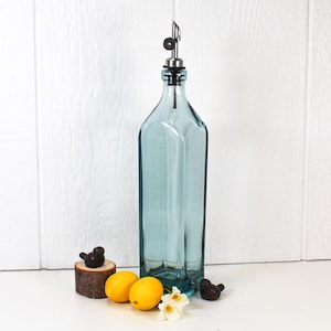 May include: A clear blue glass bottle with a silver spout and a black stopper. The bottle is sitting on a white surface with two lemons and a small wooden bird figurine.
