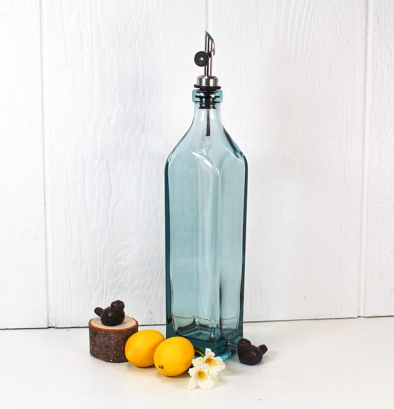 May include: A clear blue glass bottle with a silver spout and a black stopper. The bottle is sitting on a white surface with two lemons and a small wooden bird figurine.