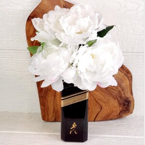 May include: White peonies in a black glass bottle with the label "Johnnie Walker Double Black" on a wooden surface.