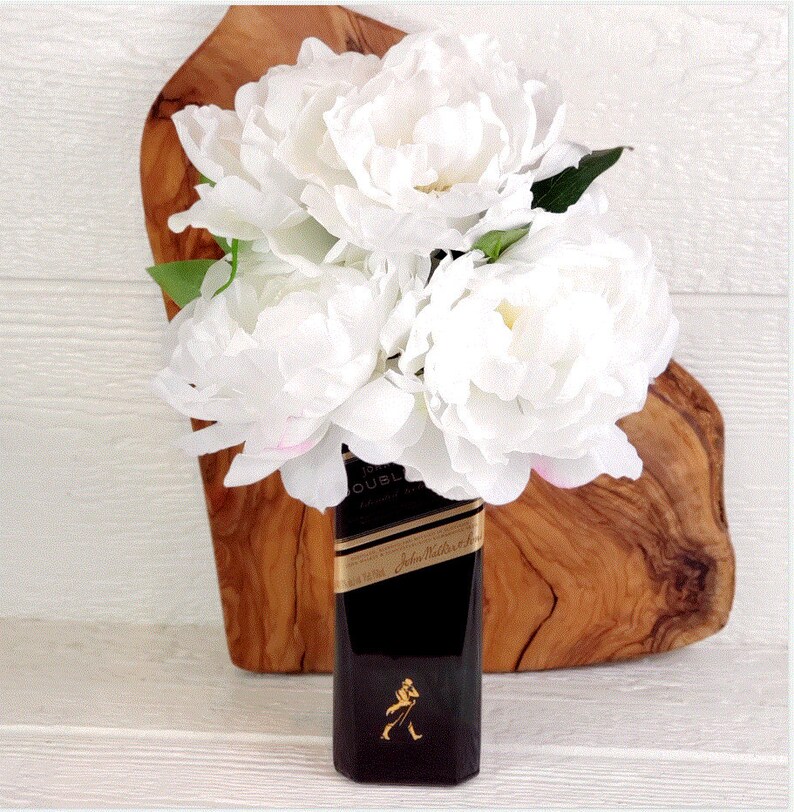 May include: White peonies in a black glass bottle with the label "Johnnie Walker Double Black" on a wooden surface.