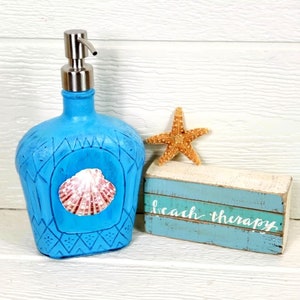May include: Blue glass soap dispenser with a seashell design and a silver pump. The dispenser is sitting on a white wooden surface with a sign that says "beach therapy".
