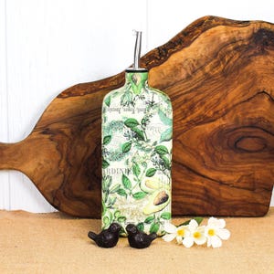 May include: A rectangular glass oil dispenser bottle featuring a botanical print with green leaves, herbs, and avocado illustrations over a light background with script text. The bottle is topped with a silver-colored metal pour spout.