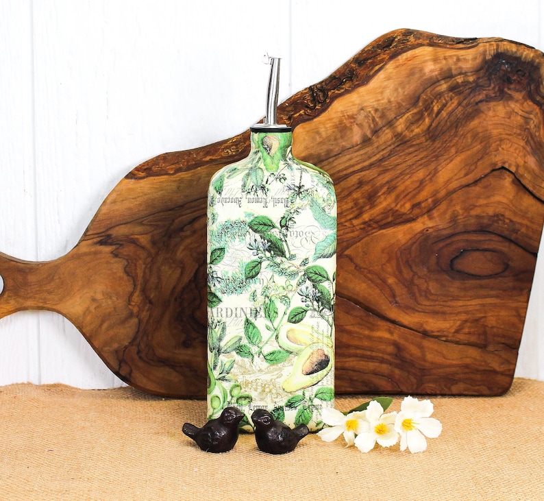 May include: A rectangular glass oil dispenser bottle featuring a botanical print with green leaves, herbs, and avocado illustrations over a light background with script text. The bottle is topped with a silver-colored metal pour spout.