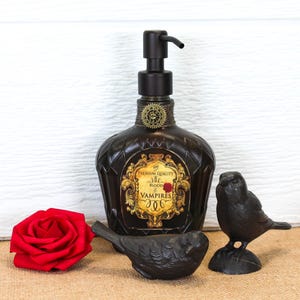 K&ouml;nnte beinhalten: Ein dunkelbrauner Seifenspender aus Glas mit schwarzer Pumpe und einem goldenen Etikett mit der Aufschrift The Premium Quality Blood of Vampires. Die Flasche ist mit einem kleinen Zahnrad verziert und wird von einer roten Rose und zwei schwarzen Vogelfiguren begleitet.