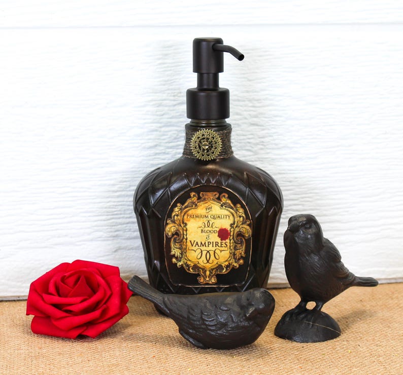 K&ouml;nnte beinhalten: Ein dunkelbrauner Seifenspender aus Glas mit schwarzer Pumpe und einem goldenen Etikett mit der Aufschrift The Premium Quality Blood of Vampires. Die Flasche ist mit einem kleinen Zahnrad verziert und wird von einer roten Rose und zwei schwarzen Vogelfiguren begleitet.