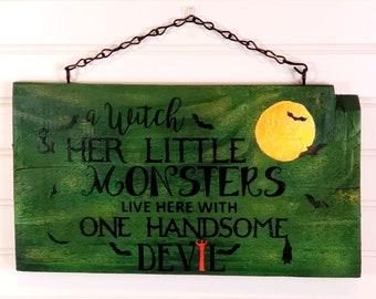 Op de afbeelding: Een groen houten Halloween-bord met zwarte tekst die luidt: "A Witch & Her Little Monsters Live Here With One Handsome Devil". Het bord heeft een gele maan, vleermuizen en een rode duivelvork. Een zwarte ketting is bevestigd om op te hangen.