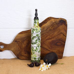 May include: A tall glass bottle with a pour spout, decorated with a green and white botanical print featuring avocado illustrations and text. The bottle is positioned next to a whole dark purple avocado and two small dark bird figurines.