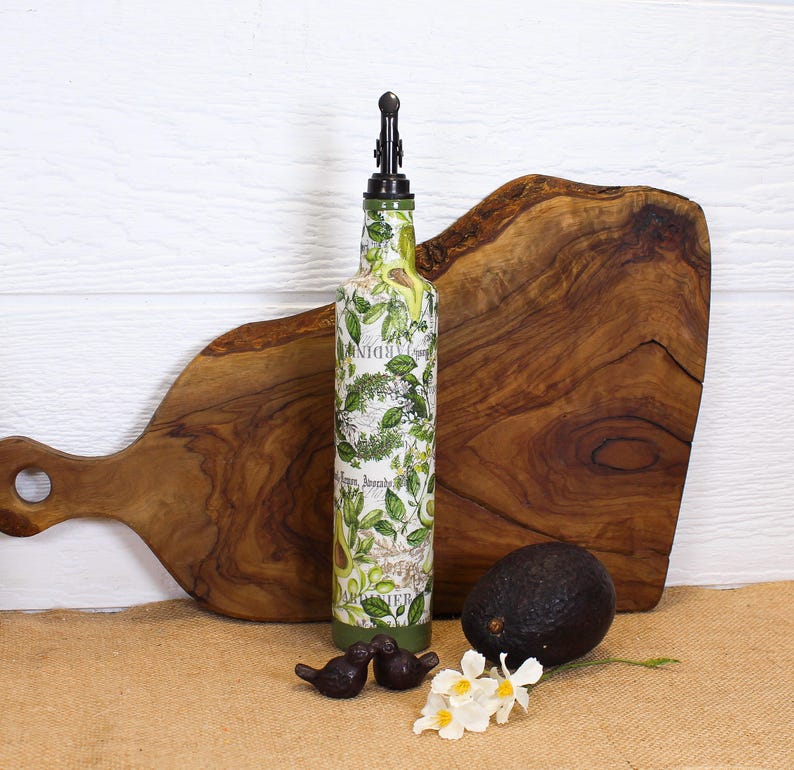 May include: A tall glass bottle with a pour spout, decorated with a green and white botanical print featuring avocado illustrations and text. The bottle is positioned next to a whole dark purple avocado and two small dark bird figurines.