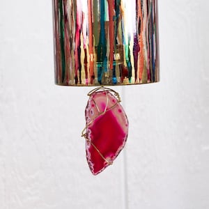 May include: A decorative wind chime featuring a cylindrical top with vertical streaks of gold, green, blue, and red. A vibrant pink and white agate slice is suspended below, wrapped in gold wire. The wind chime is designed for indoor or outdoor use.