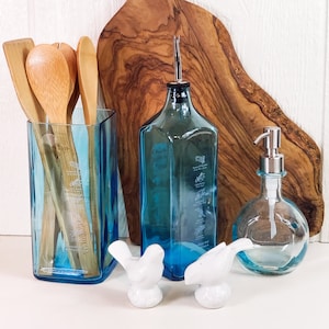 Op de afbeelding: Een blauwe glazen fles met een kurkstop, een blauwe glazen zeepdispenser en een blauwe glazen vaas met houten lepels erin. De items staan op een wit oppervlak met een houten snijplank erachter.