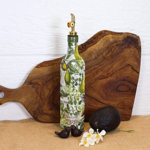 May include: A glass bottle with a gold-colored metal pourer spout, decorated with a botanical print featuring green avocados and leafy herbs on a white background. The bottle is cylindrical with a narrow neck and a green cap base for the spout.