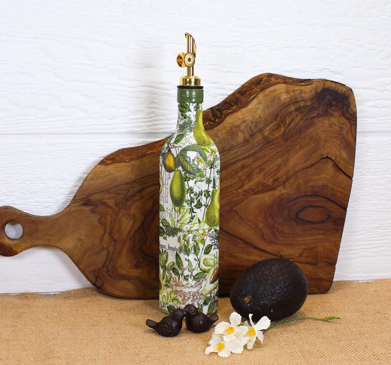 May include: A glass bottle with a gold-colored metal pourer spout, decorated with a botanical print featuring green avocados and leafy herbs on a white background. The bottle is cylindrical with a narrow neck and a green cap base for the spout.