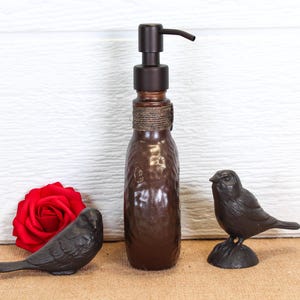 K&ouml;nnte beinhalten: Ein brauner Seifenspender aus Glas mit einem dunklen Metallpumpmechanismus und einem umwickelten Bindfaden am Hals, begleitet von zwei kleinen schwarzen Vogelfiguren und einer leuchtend roten Rose auf einer ebenen Fl&auml;che.