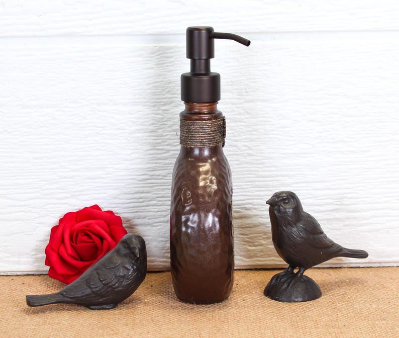 K&ouml;nnte beinhalten: Ein brauner Seifenspender aus Glas mit einem dunklen Metallpumpmechanismus und einem umwickelten Bindfaden am Hals, begleitet von zwei kleinen schwarzen Vogelfiguren und einer leuchtend roten Rose auf einer ebenen Fl&auml;che.