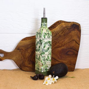 May include: A tall, square glass bottle with a silver pour spout, decorated with a green avocado and leaf pattern over a vintage-style script background. The base is painted solid green, and the bottle is positioned upright.