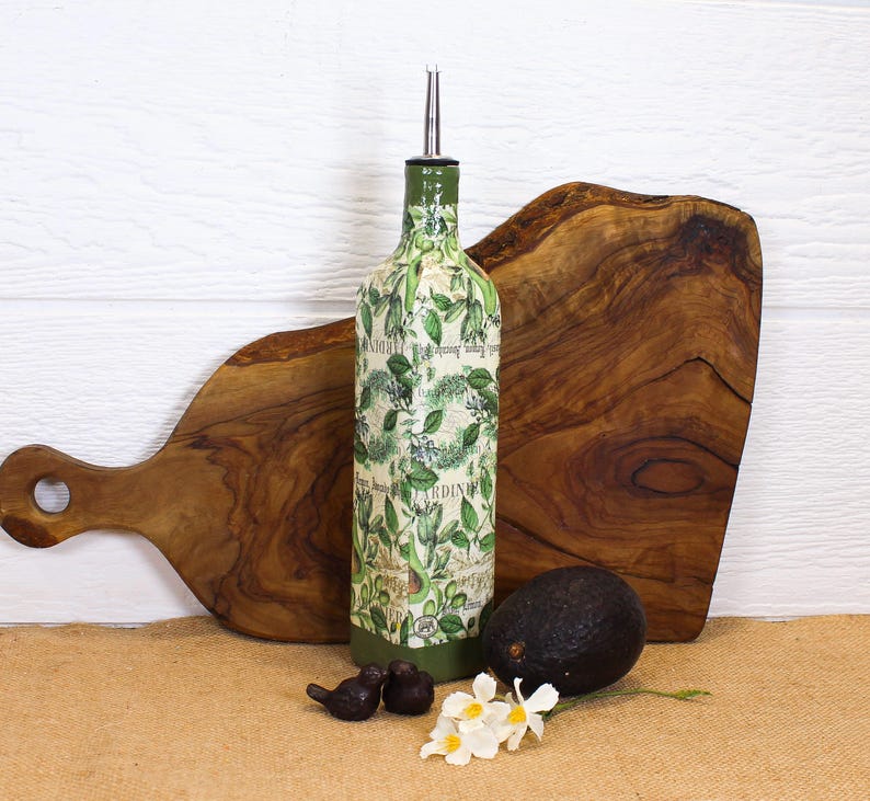 May include: A tall, square glass bottle with a silver pour spout, decorated with a green avocado and leaf pattern over a vintage-style script background. The base is painted solid green, and the bottle is positioned upright.