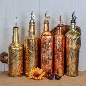 May include: Five decorative glass bottles with pour spouts. The bottles have a gold and copper metallic finish. A sunflower and two small bird figurines are in front of the bottles. The bottles are of varying shapes and sizes.