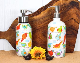May include: Two white ceramic soap dispensers with stainless steel pumps, decorated with colourful birds. A yellow sunflower and two small black bird figurines are in front of a wooden chopping board.