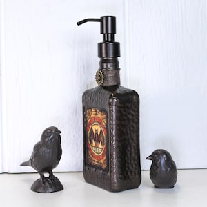 K&ouml;nnte beinhalten: Ein dunkelbrauner Seifenspender aus geh&auml;mmertem Glas mit schwarzer Pumpe, umwickelt mit Schnur und einem kleinen Zahnrad-Akzent. Das Etikett zeigt eine Fledermaus-Illustration und den Text Bat's Blood. Zwei kleine dunkle Vogelfiguren stehen daneben.