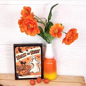 Op de afbeelding: Een houten bord met de tekst "Trick or Treat" en een illustratie van een geest en een zwarte kat. Het bord staat naast een vaas met oranje bloemen en een vaas met een candy corn-thema.