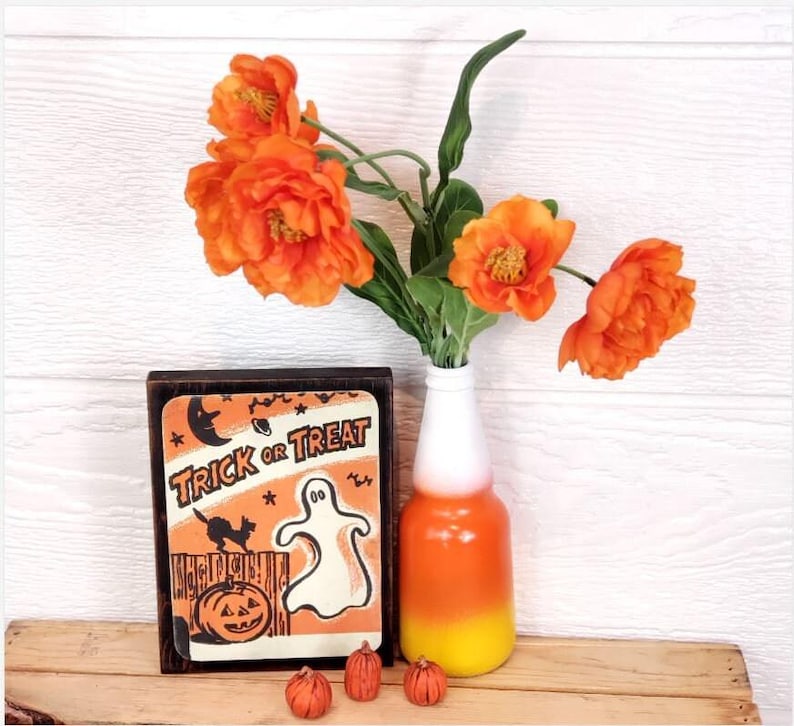 Op de afbeelding: Een houten bord met de tekst "Trick or Treat" en een illustratie van een geest en een zwarte kat. Het bord staat naast een vaas met oranje bloemen en een vaas met een candy corn-thema.