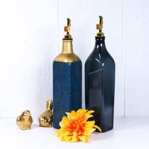 May include: Two glass bottles with gold and blue paint, one with a gold top and one with a black top, on a white surface with a yellow flower and two small bird figurines.