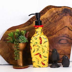 May include: A yellow and red ceramic soap dispenser with a black pump, decorated with a pattern of red and green chili peppers. The dispenser is sitting on a wooden cutting board with a small potted succulent plant and two black metal bird figurines.