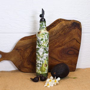 May include: A tall, cylindrical glass oil dispenser bottle featuring a botanical print of green avocados, leaves, and herbs with the text 'Botanical' and 'Basil' printed on the surface. The bottle is topped with a black plastic pour spout.