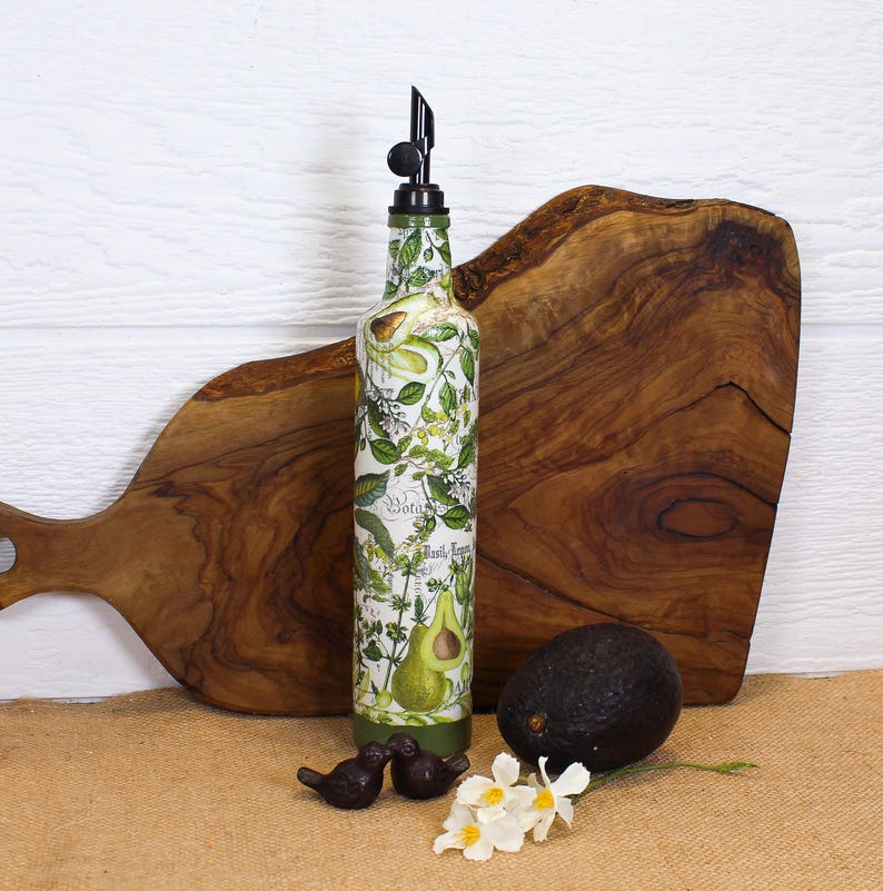 May include: A tall, cylindrical glass oil dispenser bottle featuring a botanical print of green avocados, leaves, and herbs with the text 'Botanical' and 'Basil' printed on the surface. The bottle is topped with a black plastic pour spout.