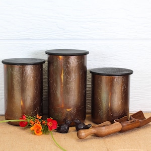 May include: Three cylindrical canisters with a textured, metallic bronze finish and flat black lids. Two small black bird figurines and two wooden scoops are placed in front of the canisters, alongside a small cluster of orange and red flowers.