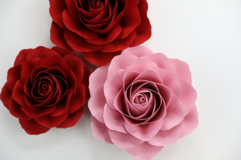 Puede incluir: Tres rosas de papel, una roja, una rosa y una rojo oscuro, est&aacute;n dispuestas sobre un fondo blanco. Las rosas est&aacute;n hechas de papel y tienen un aspecto tridimensional.