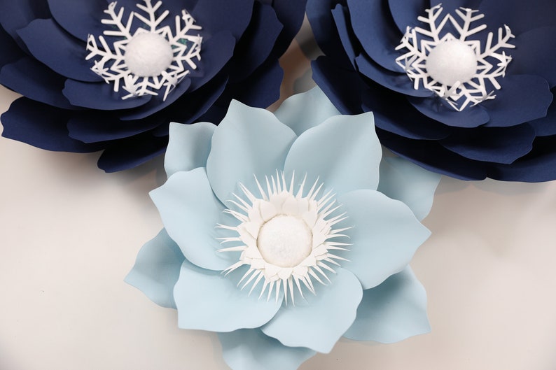 Pu&ograve; includere: Tre fiori di carta, uno azzurro chiaro e due blu navy, con centri a fiocco di neve bianco. I fiori sono sovrapposti su uno sfondo bianco.