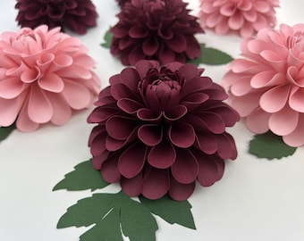 May include: Close-up of several paper dahlias in shades of burgundy and pink, with green paper leaves. The flowers are arranged on a white surface, showcasing their layered petal design. These handcrafted paper flowers are ideal for home decor or event decorations.