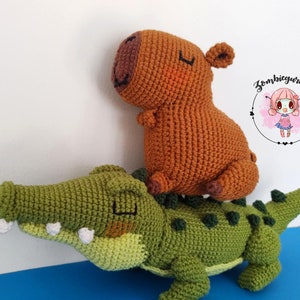Puede incluir: Un cocodrilo de crochet verde con un capibara de crochet marrón sentado en su espalda. El cocodrilo tiene una sonrisa de dientes y el capibara tiene una expresión dulce.