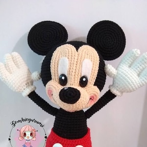 Puede incluir: Un juguete de peluche Mickey Mouse tejido a crochet con orejas negras, cuerpo negro, pantalones cortos rojos con botones blancos, zapatos amarillos y guantes blancos. El juguete está de pie con los brazos extendidos.