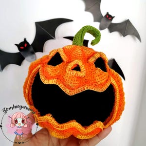 Puede incluir: Una calabaza naranja tejida a ganchillo con una cara tallada y un tallo verde. La calabaza tiene una sonrisa amplia y con dientes y ojos triangulares. Murciélagos de papel negro con ojos rojos en el fondo. Decoración de Halloween.