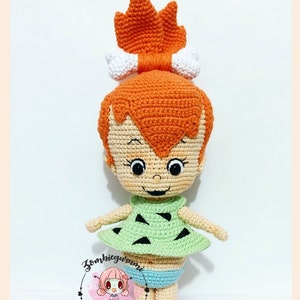May include: A crocheted doll of a cartoon character with orange hair, a white bone in her hair, a green dress with black triangles, and blue underwear. The doll is standing on a white surface.