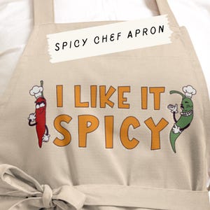 Puede incluir: Delantal de chef beige con el texto "I LIKE IT SPICY" en letras naranjas. El delantal presenta dibujos animados de chiles con gorros de chef. La parte superior del delantal tiene el texto "SPICY CHEF APRON".