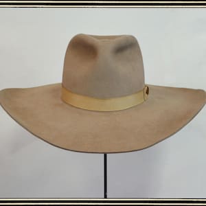 Pode incluir: Chapéu bege de abas largas com uma faixa castanha clara. O chapéu tem um formato fedora clássico com uma coroa ligeiramente côncava. A aba é larga e suavemente curvada, exibido em um suporte preto.