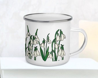 May include: A white enamel mug with a silver rim and a handle, featuring a repeating illustration of white snowdrop flowers with green stems and leaves wrapped around the exterior surface of the cup.