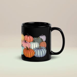 May include: A black ceramic mug featuring a colorful illustration of various pumpkins in shades of orange, pink, blue, white, and green. The design displays a cluster of pumpkins with distinct ribbed textures and stems, arranged in a dense, overlapping pattern.
