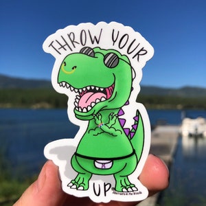 May include: A green dinosaur sticker with sunglasses and a purple tail. The sticker says "Throw Your Up" and has the text "Abstracts in the Woods" at the bottom.