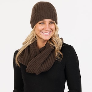 May include: A brown ribbed knit beanie hat and infinity scarf. The hat is a close-fitting style, and the scarf is wrapped around the neck. The set is made of a soft, warm material, ideal for cold weather. The woman is wearing a black long-sleeve top.