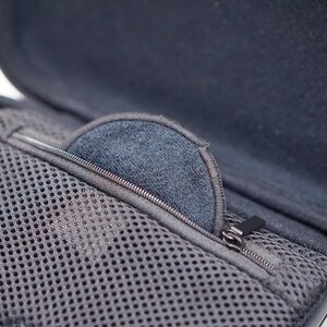 Pode incluir: Uma caixa preta com z&iacute;per, um bolso de malha e um forro macio cinza. A caixa est&aacute; aberta e um console de videogame cinza &eacute; vis&iacute;vel no fundo.