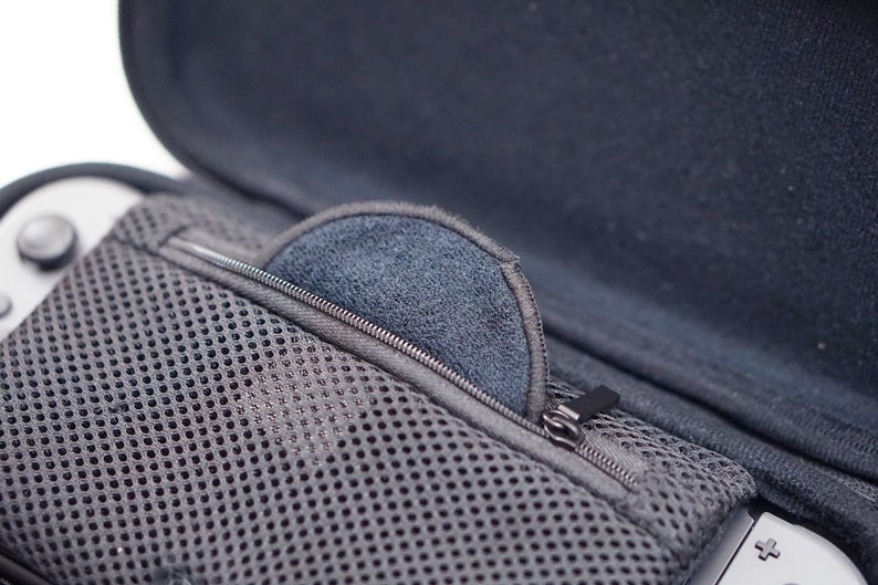 Pode incluir: Uma caixa preta com z&iacute;per, um bolso de malha e um forro macio cinza. A caixa est&aacute; aberta e um console de videogame cinza &eacute; vis&iacute;vel no fundo.