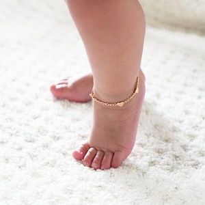 Peut inclure: Un bracelet de cheville en or avec de petits pendentifs en forme de cœur sur la cheville d'un enfant.