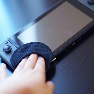 Pode incluir: Uma pessoa limpando a tela de um console Nintendo Switch preto com um pano de microfibra preto.
