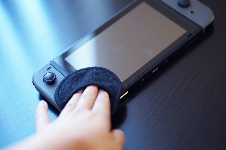Pode incluir: Uma pessoa limpando a tela de um console Nintendo Switch preto com um pano de microfibra preto.